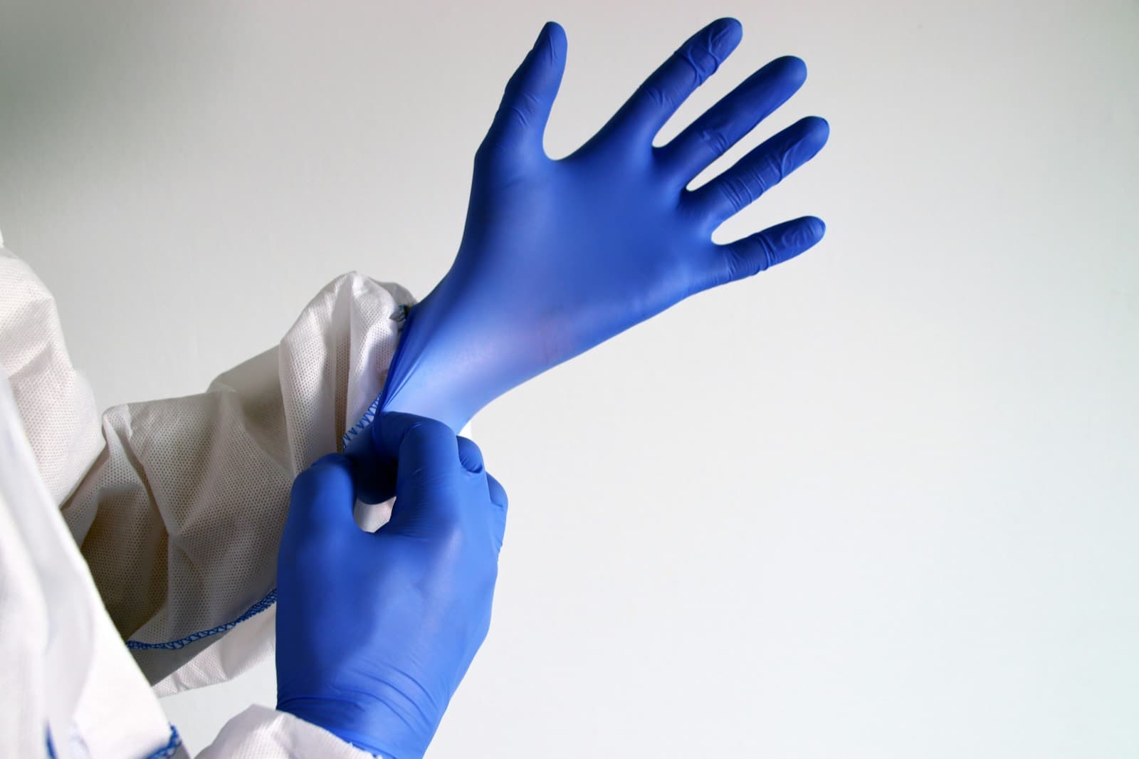 A healthcare worker in protective clothing pulling on a blue nitrile examination glove.